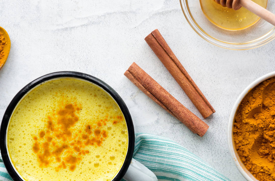 A spiced golden cacao latte in a mug next to cinnamon two sticks, honey and a bowl of cacao powder on a marble kitchen surface.