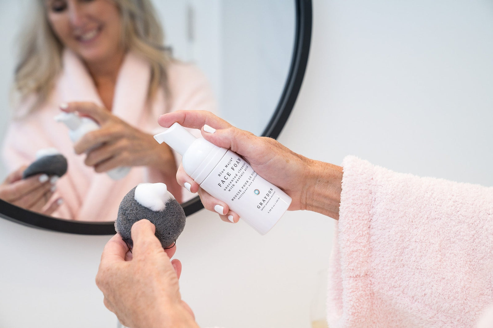 woman in fluffy robe using foaming facial cleanser, face foam, and a gently exfoliating bamboo charcoal sponge to cleanse her face