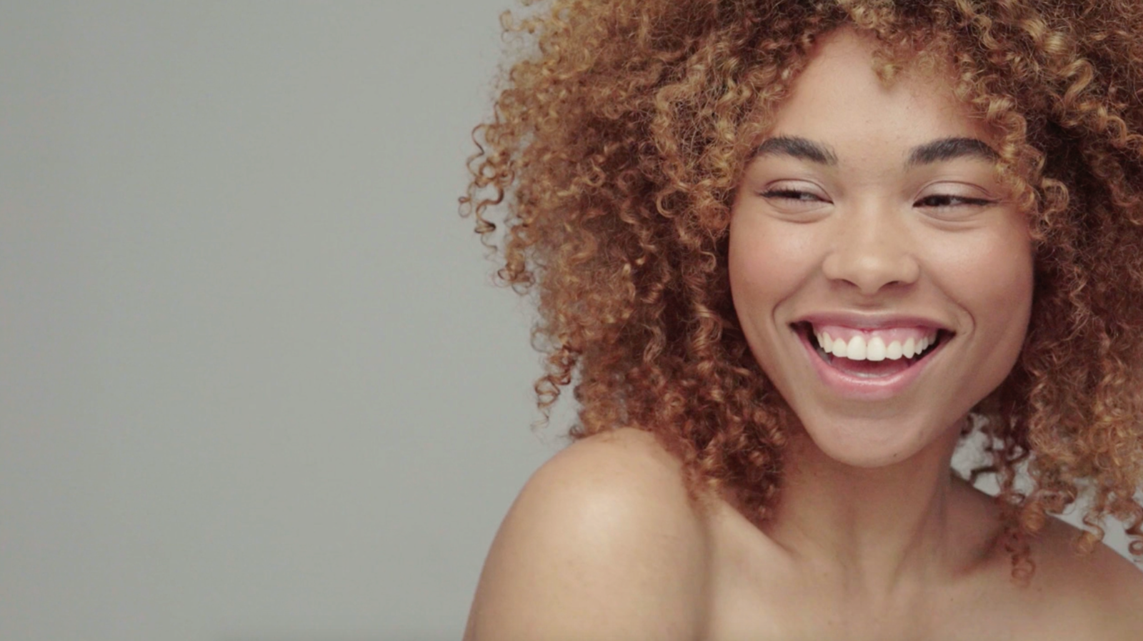 A closeup image of a dark-skinned smiling woman with shoulder-length curly hair and healthy skin 